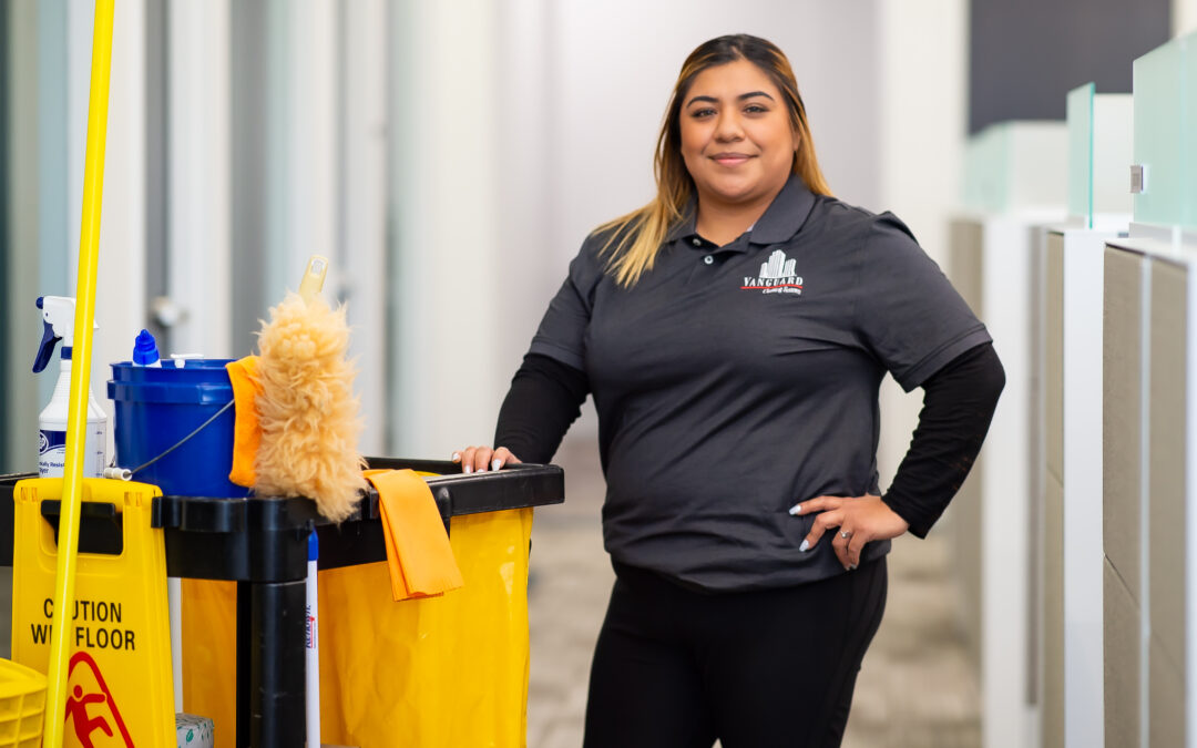 A smiling professional janitorial worker in uniform stands confidently beside a commercial cleaning cart in a modern office hallway. The cart includes cleaning supplies and a caution sign, representing high-quality, franchise-based commercial cleaning services
