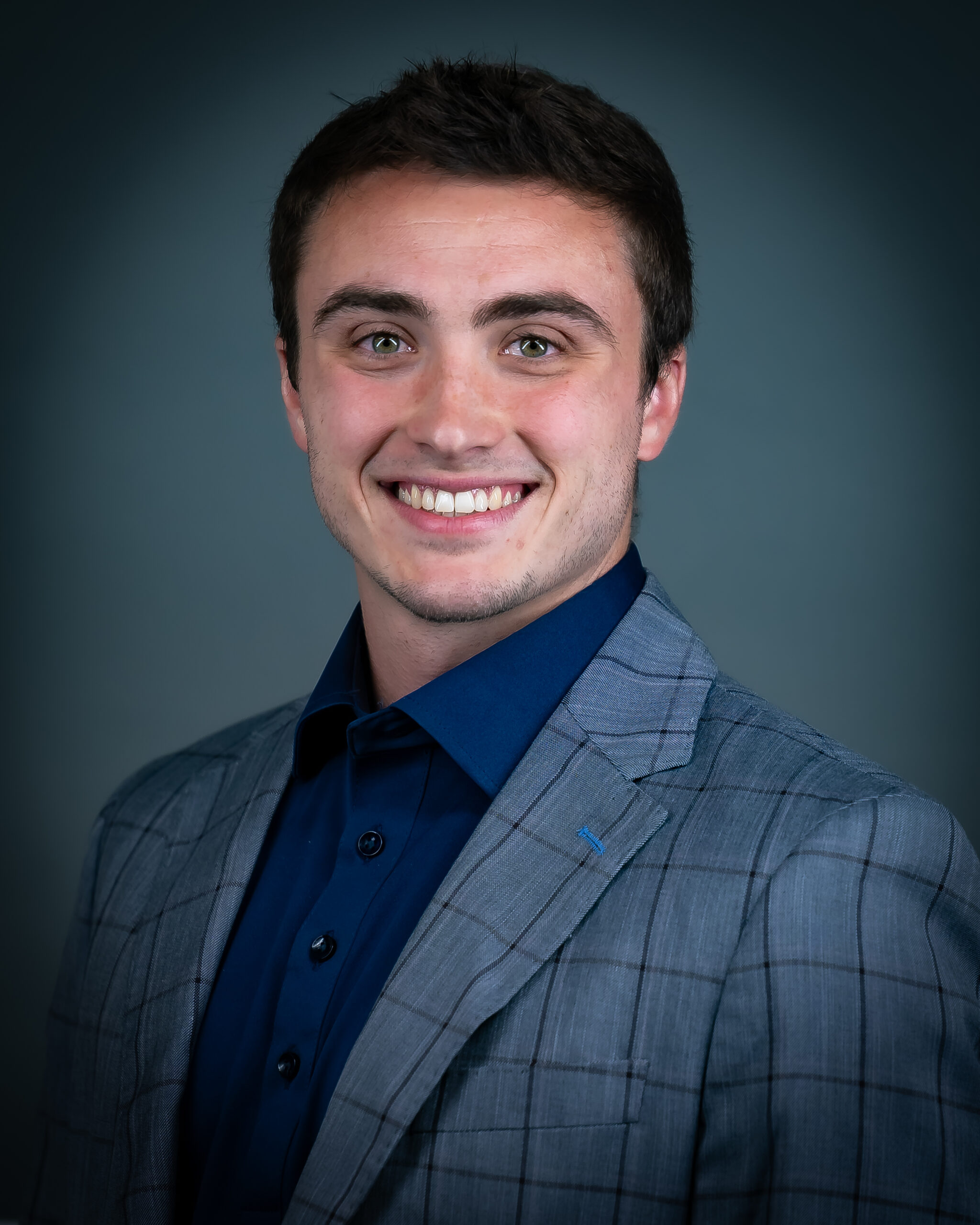 Alec Schimmel, vanguard teammate, smiling in a slate blue suit with a navy blue dress shirt. 